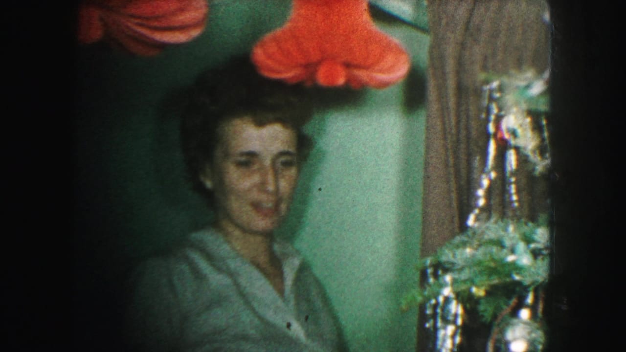 In 1959 Kansas City home-movie footage, a woman faces the camera beside a decorated Christmas tree with red holiday decor above.