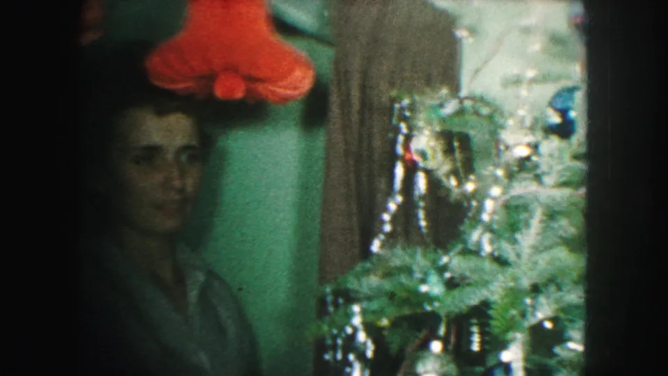 A dim 8mm frame shows the woman beside the tinsel-covered Christmas tree, with red holiday decor above her.