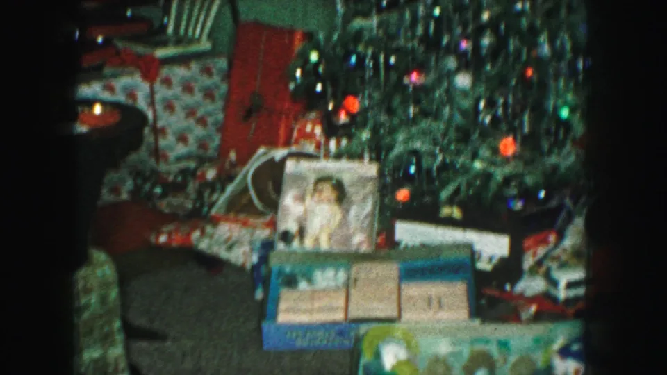 1959 Kansas City home-movie frame showing presents, toy boxes, a doll package, and tinsel on a Christmas tree in a private interior.