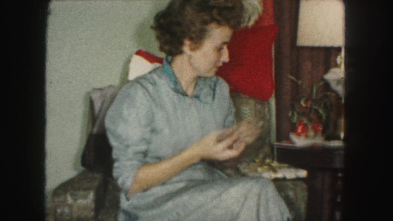 In a 1959 Kansas City home interior, a woman in a blue dress sits near an upholstered chair, side table, lamp, and Christmas stocking.
