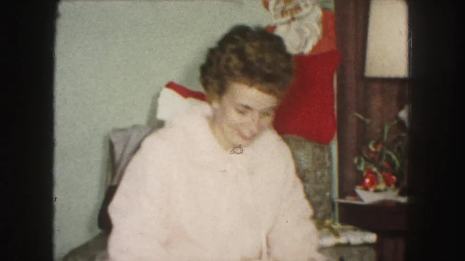 A woman in a pale pink robe sits in a 1959 Kansas City living room with a Christmas stocking, lamp, and side table behind her.