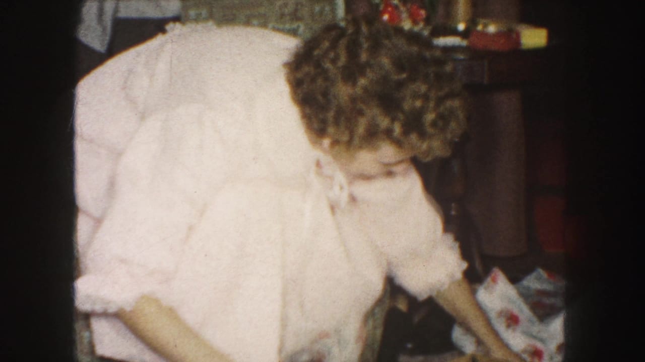 The woman in a pink robe bends toward loose wrapping paper in a private 1959 Kansas City holiday interior.