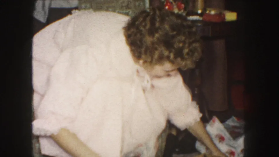 The woman in a pink robe bends toward loose wrapping paper in a private 1959 Kansas City holiday interior.