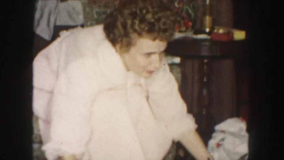 A woman in a pink robe reaches downward near loose paper in a 1959 Kansas City home decorated for Christmas.
