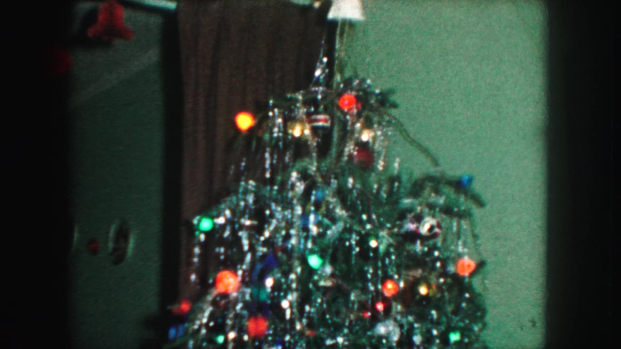 Lit Christmas tree with tinsel, colored bulbs, and an angel topper near a curtain.