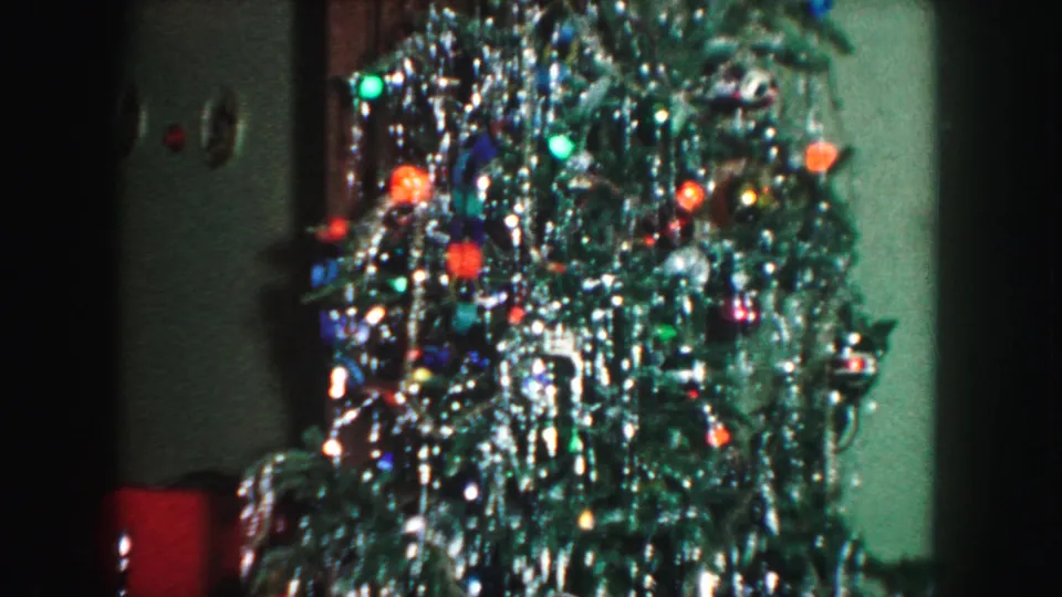 1959 Kansas City frame showing the lit Christmas tree in a dim room, with tinsel, colored bulbs, green wall, and curtain behind it.