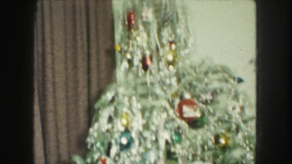 Close 1959 home-movie view of a tinsel-heavy Christmas tree with colorful ornaments against brown curtains.