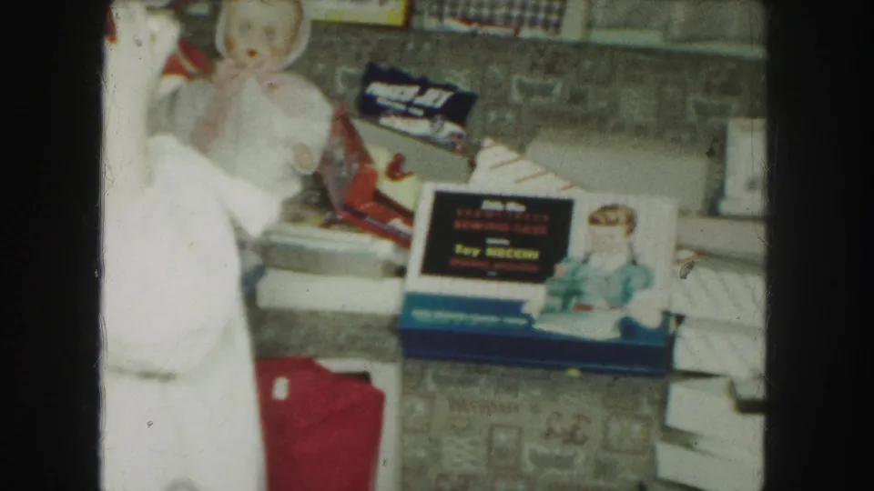 1959 Kansas City home-movie frame showing a white nightgown at left beside dolls, a blue toy box, scattered packaging, and gift boxes.
