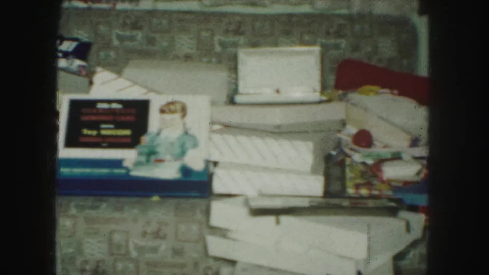 1959 Kansas City frame with a doll beside a blue toy box, open packaging, white boxes, and holiday gifts on patterned carpet.