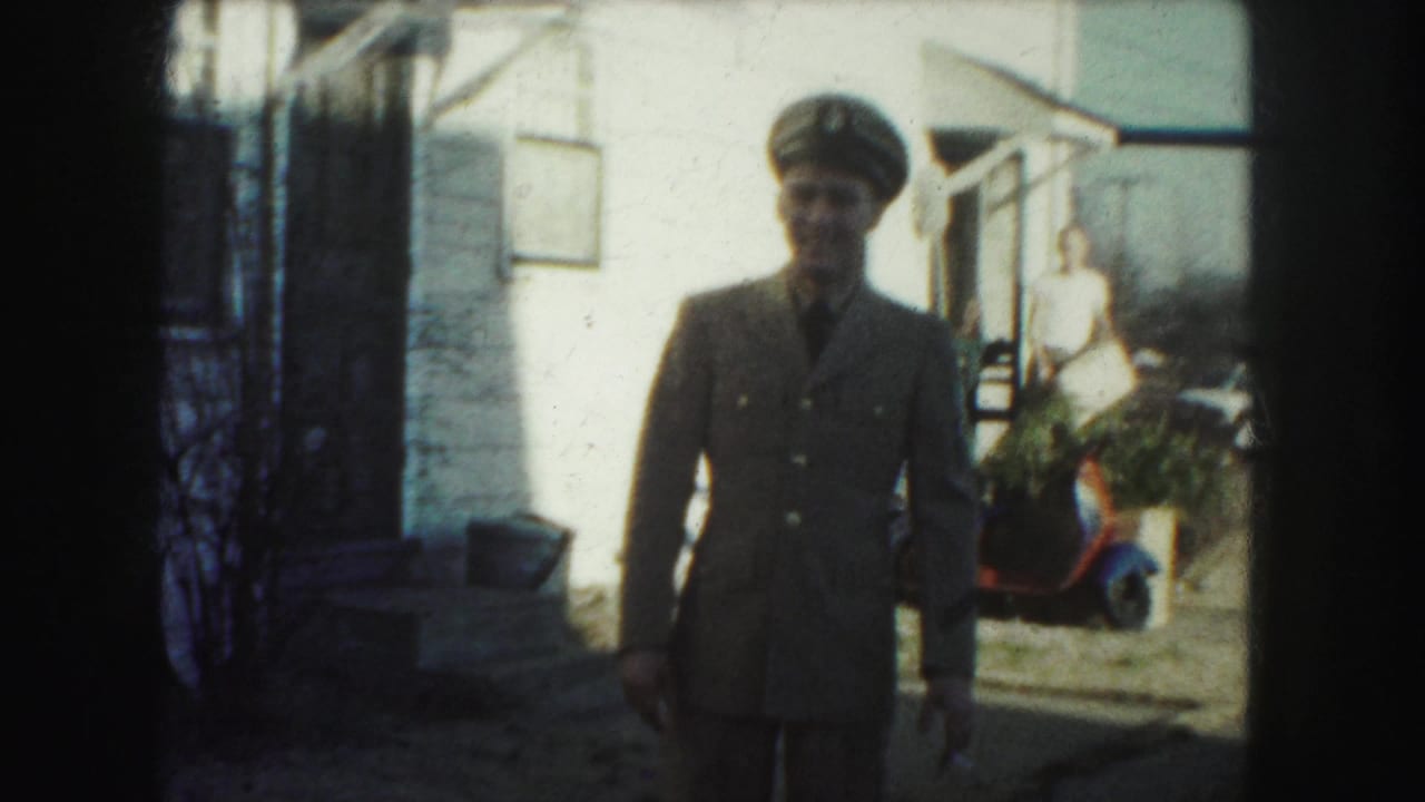 Uniformed man in a peaked cap stands in a yard near a white house.