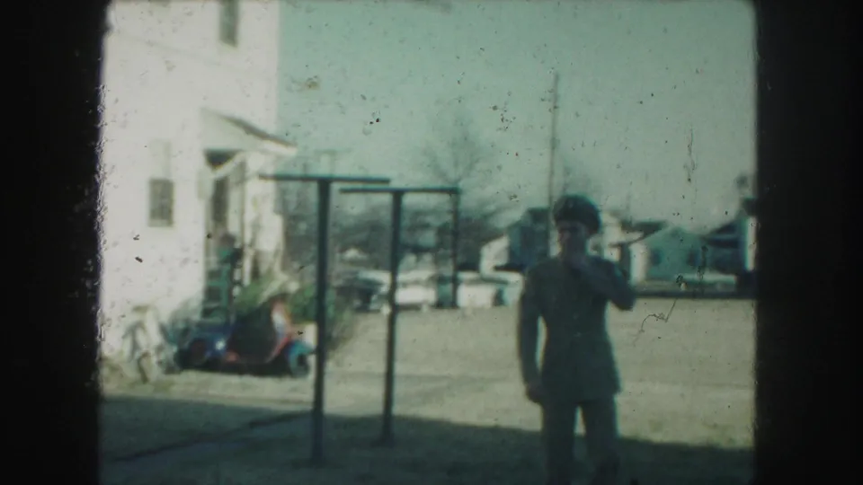 The uniformed man stands in the residential yard and raises a hand near his face, with a white house and scooter behind him.