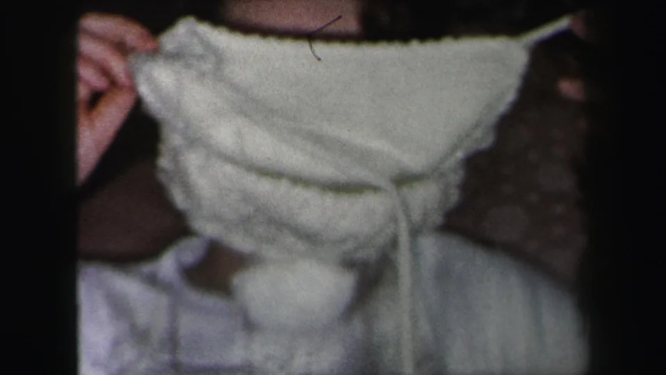 1975 Lynbrook 8mm frame showing a young woman using both hands to adjust a soft white knitted hat over her face.