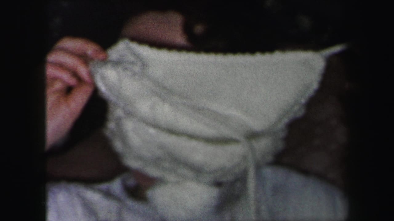 In 1975 Lynbrook home-movie footage, a young woman in white holds a bonnet or fabric over her face while seated indoors.