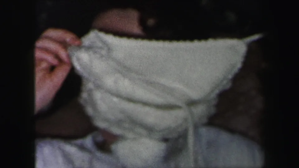 In 1975 Lynbrook home-movie footage, a young woman in white holds a bonnet or fabric over her face while seated indoors.
