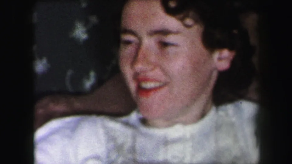 A close 1975 Lynbrook home-movie frame shows the young woman smiling and turning slightly in a white garment indoors.