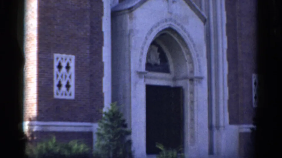 1975 Bridgeport home-movie frame showing a brick church exterior with a pale stone arched entrance and visible 8mm gate edges.