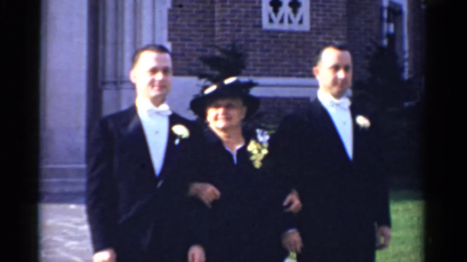 1975 Bridgeport wedding home movie showing two men in black tuxedos posing with an older woman in a feathered hat outside a brick church.