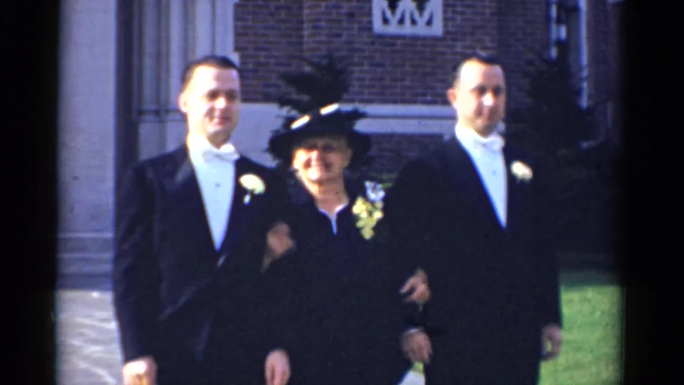 1975 Bridgeport frame of three adults in wedding attire, including two tuxedoed men and an older woman in a black hat, posing outside church.