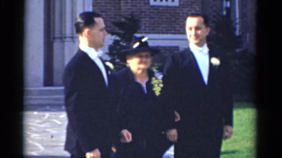 In 1975 Bridgeport wedding footage, two tuxedoed men stand with a formally dressed woman outside a brick building entrance.