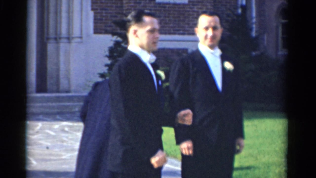 Two men in tuxedos walk past a woman near a building entrance and lawn.