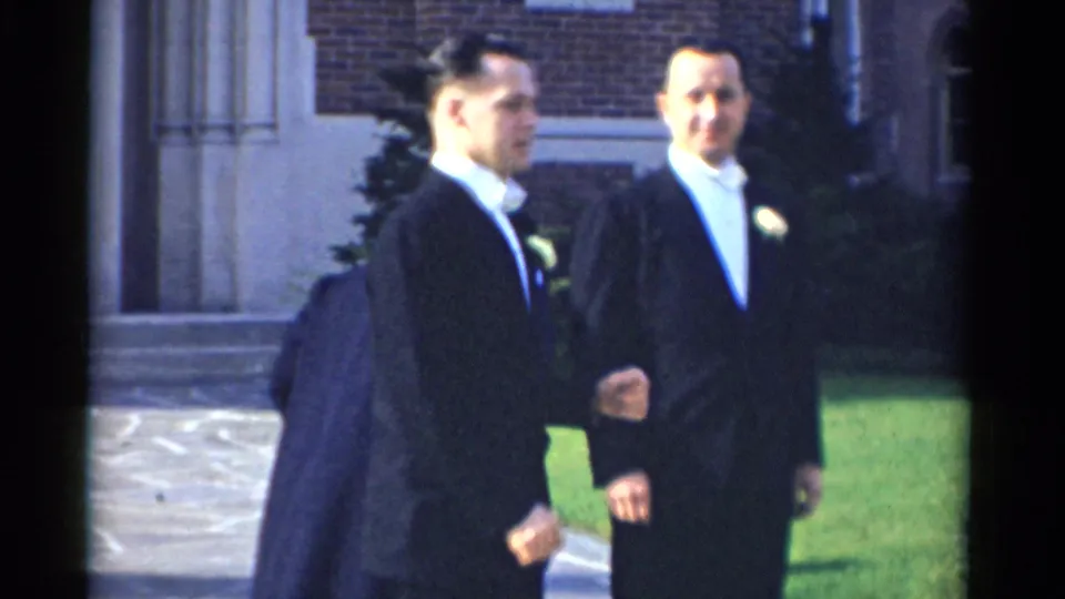 Two tuxedoed men walk outside during the Bridgeport wedding scene, with a woman partly visible near the brick building and lawn.