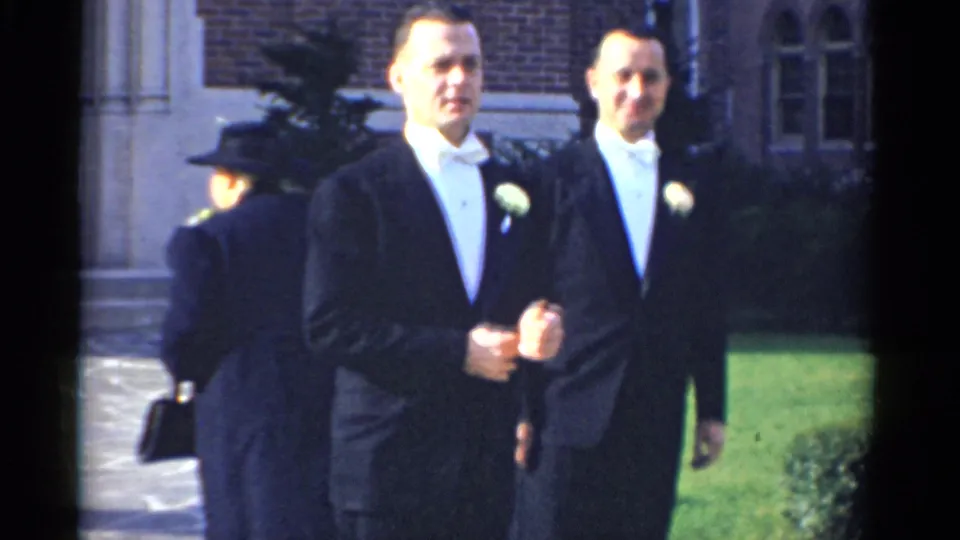 A man in a black tuxedo and white bow tie walks toward the home-movie camera, with another tuxedoed man behind him.