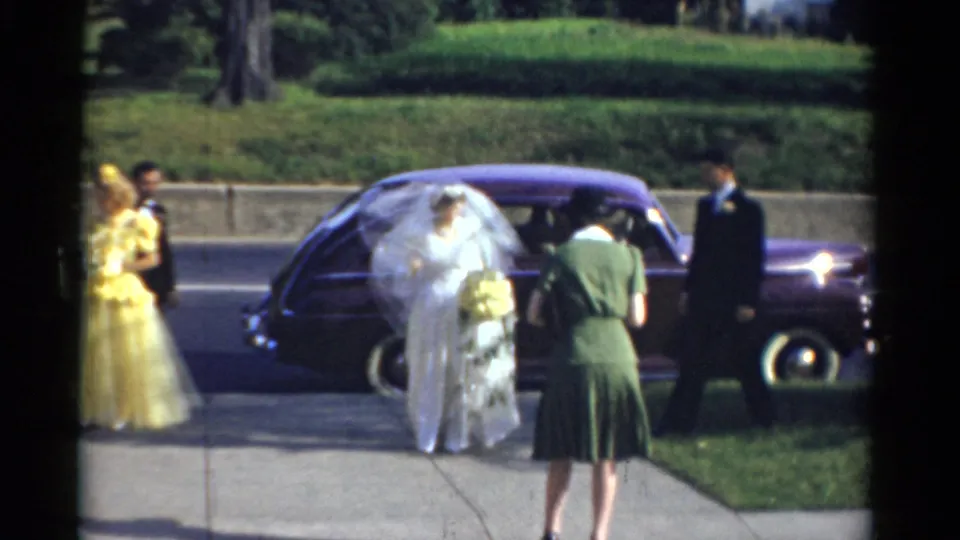 The bride arrives beside a dark car in 1975 Bridgeport wedding footage, holding flowers while guests and a yellow-dressed attendant stand nearby.