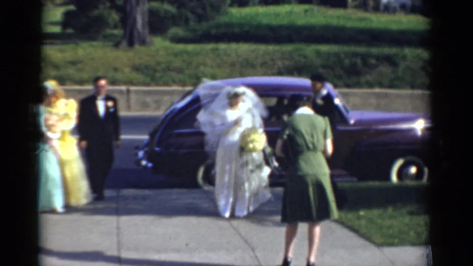 In Bridgeport, Connecticut, 1975 wedding footage, a veiled bride with bouquet and groom stand near a purple car as attendants gather nearby.