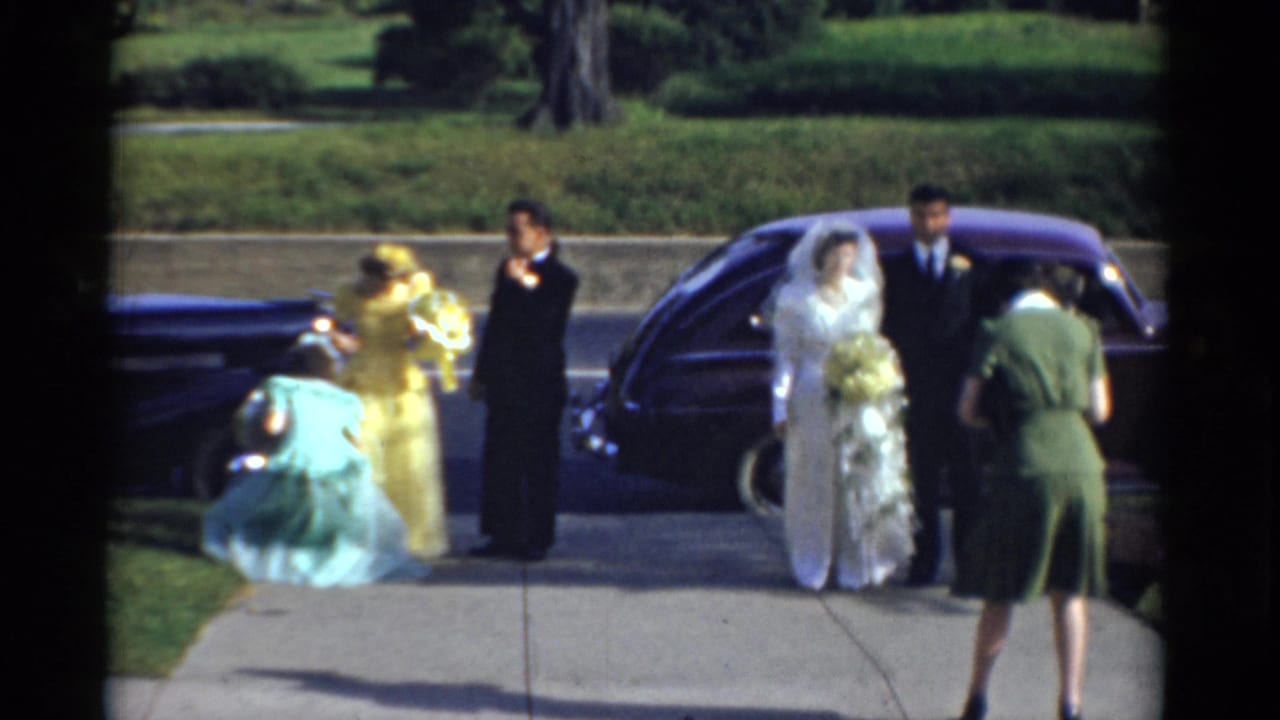 The bride and groom stand with attendants near the purple car while a turquoise-dressed guest bends down in the foreground.
