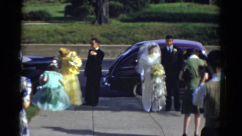 A veiled bride holding flowers stands beside the groom and a dark-suited man as guests cluster near the sidewalk and parked car.