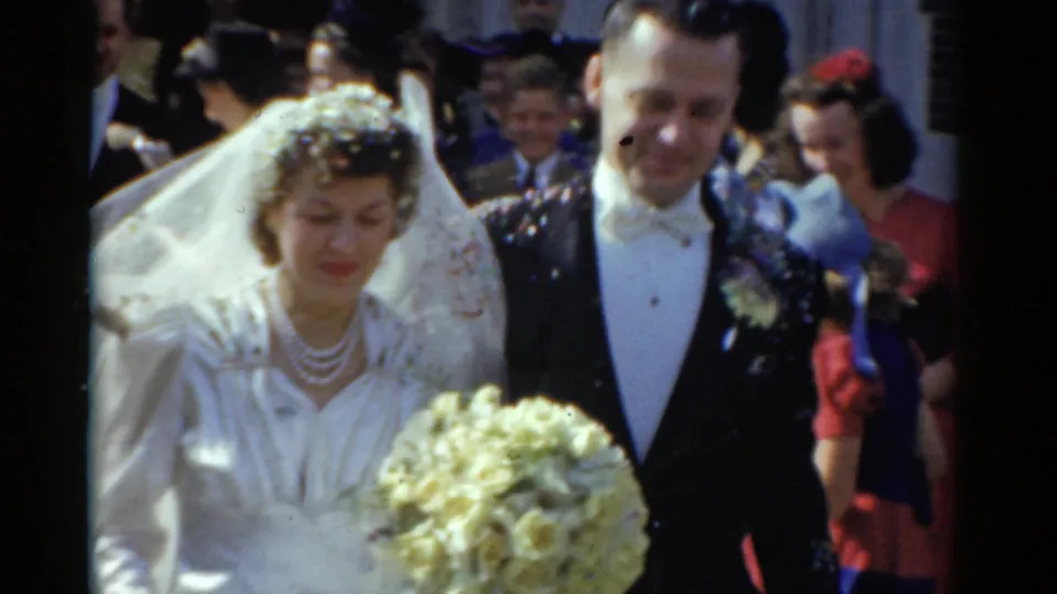 The 1975 Bridgeport bride and groom move past the camera after the church exit, with veil, bouquet, tuxedo, and guests behind them.
