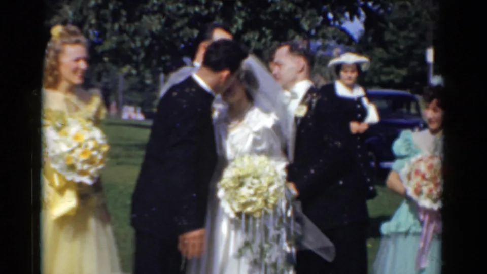 Outdoor 1975 Bridgeport wedding moment as the groom kisses the veiled bride beside attendants, guests, lawn, and a dark car.