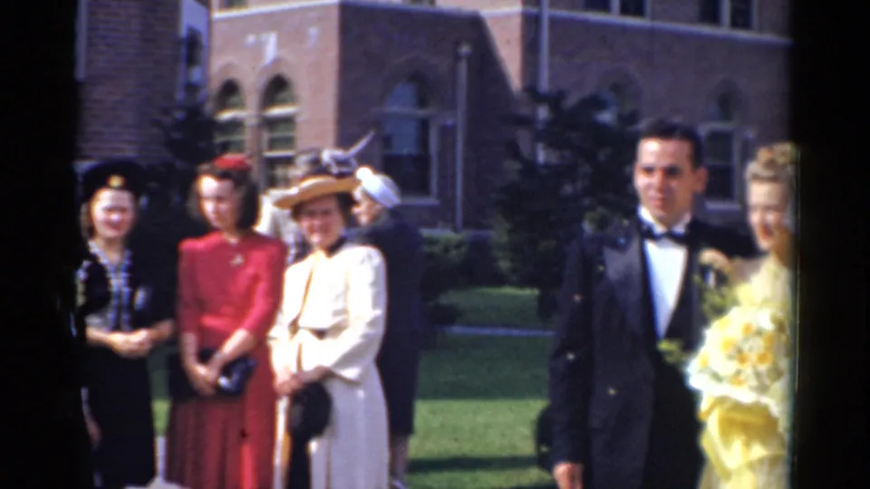 A groom in a tuxedo stands near guests and a woman with a bouquet outside the church in 1975 Bridgeport wedding footage.