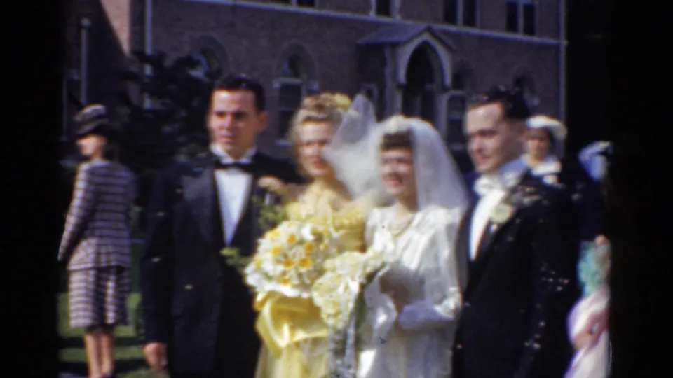 Bride, groom, bridesmaid with bouquet, and another tuxedoed man stand together outside the church in 1975 Bridgeport footage.