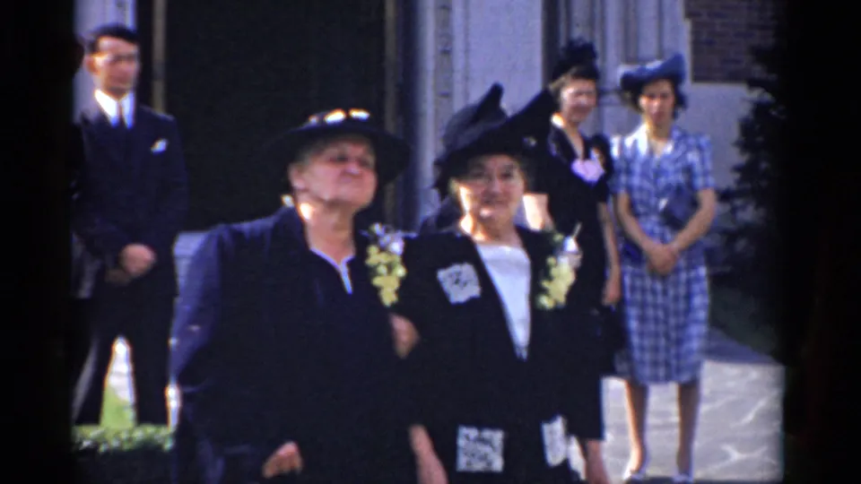 Two women in dark formal outfits with corsages walk arm-in-arm outside a Bridgeport brick building in 1975 home-movie footage.