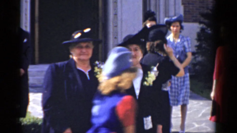 Formal guests in hats and dresses gather near a walkway outside a Bridgeport brick building, with handheld 1975 8mm framing.