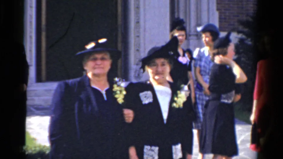 Two women in dark formal outfits and corsages face the camera outside a Bridgeport building, with other formally dressed guests behind them.