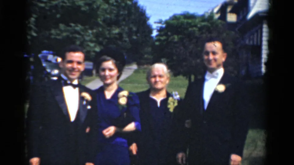 Bridgeport 1975 frame of formally dressed adults with boutonnieres and corsages standing on a lawn beside trees and a house edge.