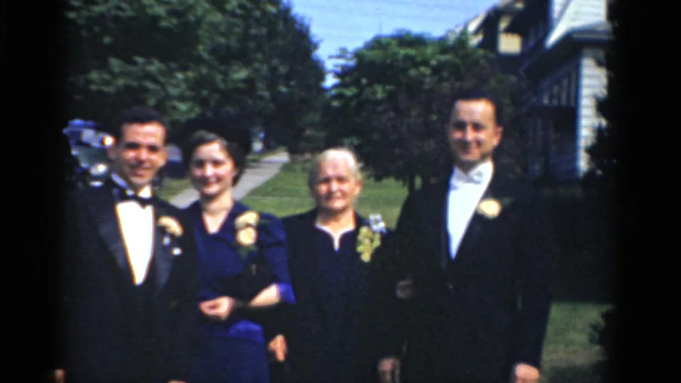 Bridgeport 1975 home-movie frame showing a group in formal wear posing outside near a sidewalk, trees, parked cars, and a house.