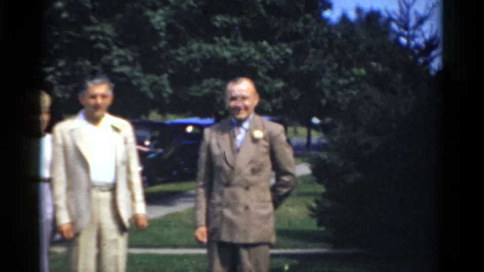 Bridgeport 1975 frame showing two older men, one in a light suit and one in a brown double-breasted suit, standing outdoors on a lawn.