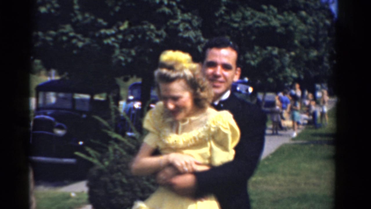 Tuxedoed man embraces a woman in a yellow formal dress outdoors near cars and trees.
