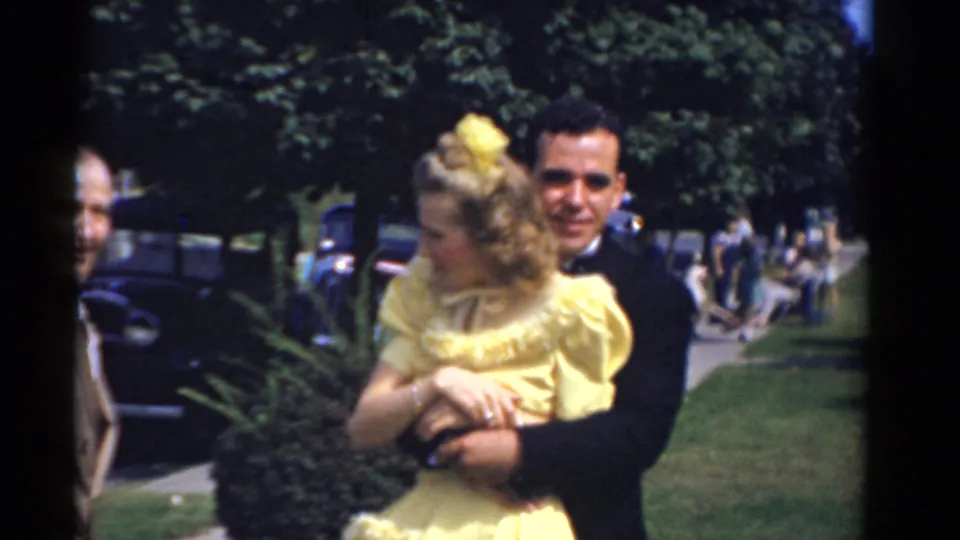 A woman in a yellow formal dress turns toward the side while a tuxedoed man embraces her in outdoor 1975 Bridgeport wedding footage.
