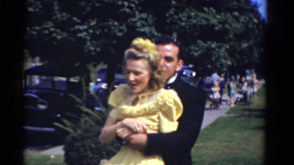 The couple stands outdoors during the Bridgeport wedding, with the woman in yellow framed by the tuxedoed man, cars, grass, and trees.