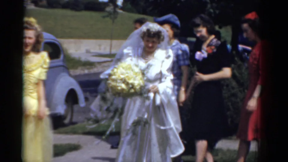 The bride walks outdoors in a white gown with a yellow bouquet, accompanied by attendants in yellow, blue, black, and red dresses.