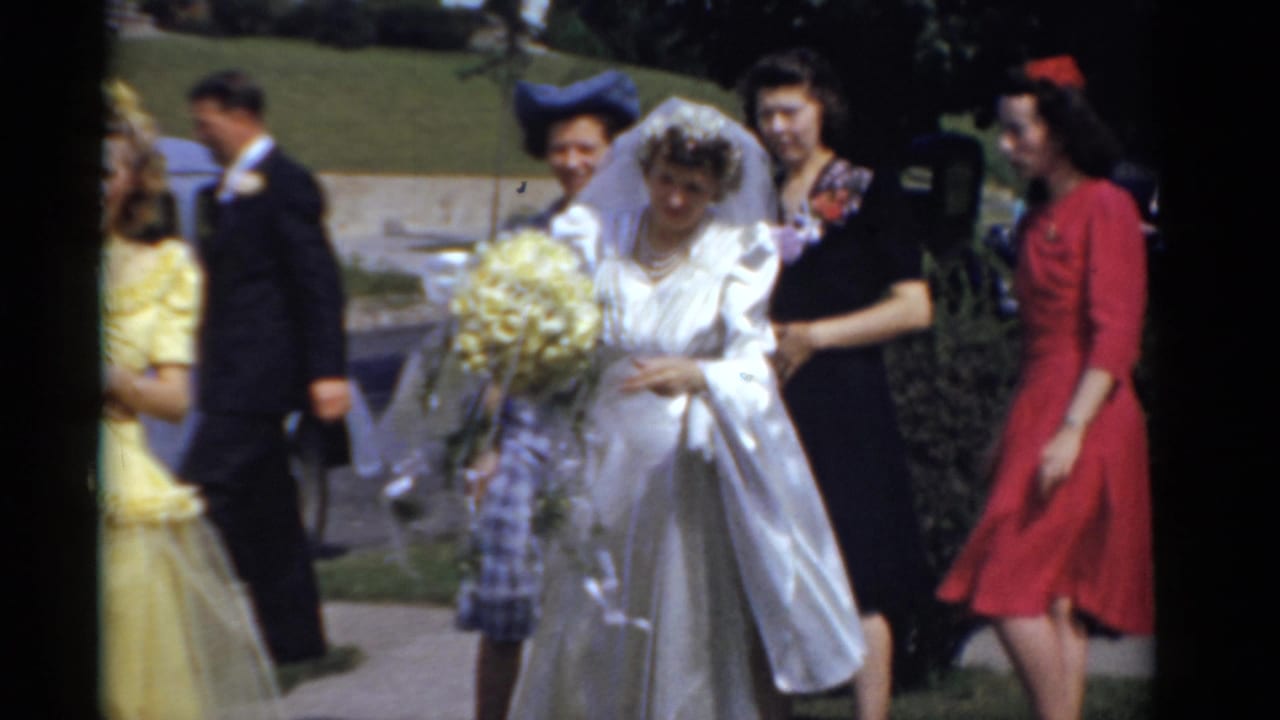 1975 Bridgeport wedding home-movie frame with bride holding yellow bouquet as bridesmaids walk nearby on a sunny sidewalk.
