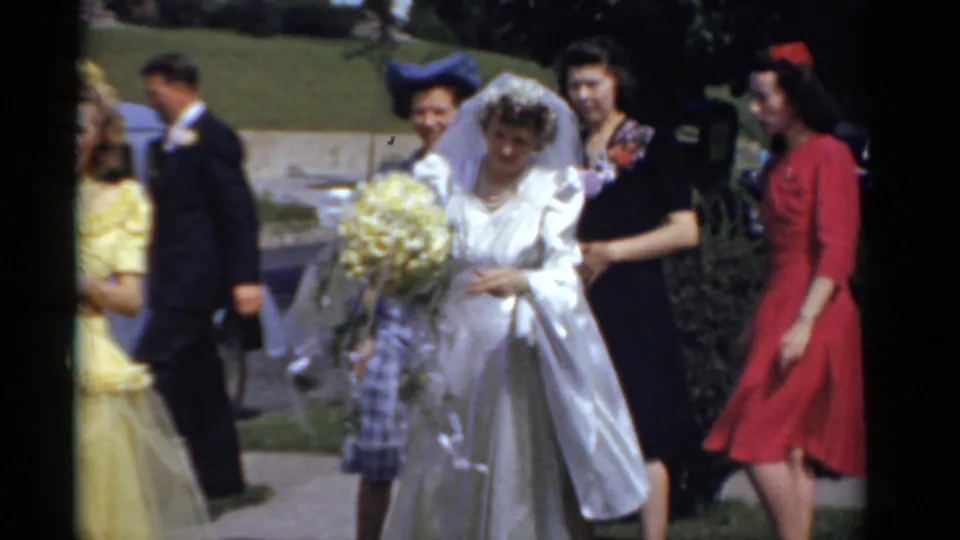 1975 Bridgeport wedding home-movie frame with bride holding yellow bouquet as bridesmaids walk nearby on a sunny sidewalk.