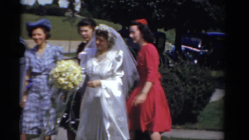 Bride in a white satin gown walks with attendants in red and patterned dresses in 1975 Bridgeport home-movie footage.