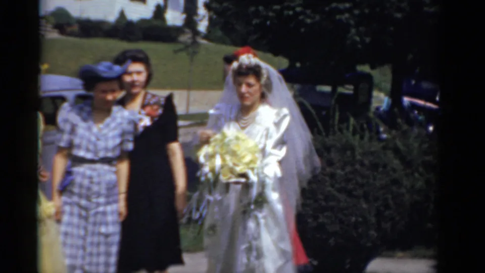Outdoor 1975 wedding frame showing the bride and attendants moving past greenery with classic cars in the background.