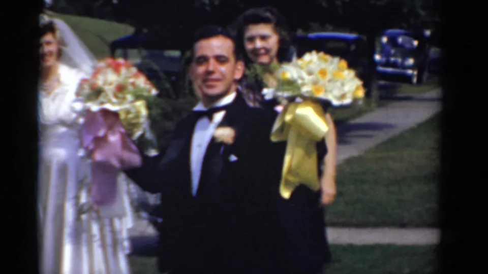 A tuxedoed man, likely the groom, smiles toward the camera while holding bouquets during a 1975 Bridgeport wedding scene.