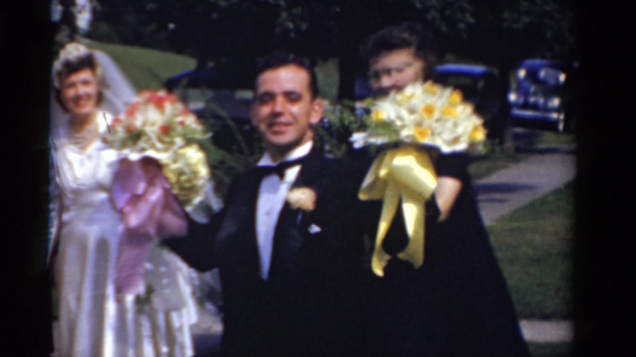 In 1975 Bridgeport wedding footage, the groom stands beside the bride and bridesmaids holding ribboned bouquets outdoors.